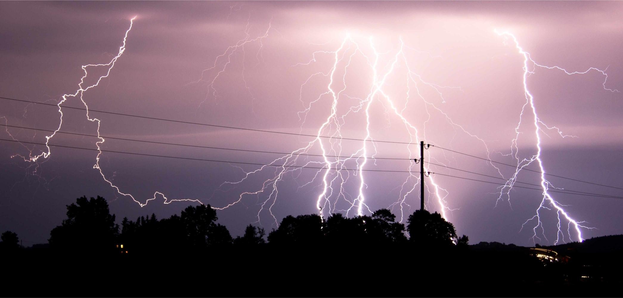 Ein nächtlicher Himmel wird dramatisch von zahlreichen grellen Blitzen durchzogen, die sich verzweigend über die gesamte Bildfläche erstrecken. Die elektrische Entladung taucht die Szene in ein violett-weißes Licht. Im Vordergrund zeichnen sich die dunklen Silhouetten von Bäumen und einem Strommast mit Überlandleitungen ab, die sich quer durch das Bild ziehen. Die Lichtspuren und die dichte Struktur der Blitze lassen die enorme Energie des Gewitters erahnen und vermitteln eine zugleich bedrohliche wie beeindruckende Atmosphäre.