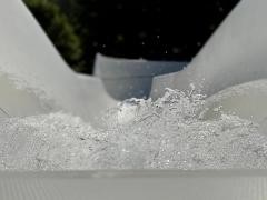 Eine weiße Wasserrutsche endet in einem spritzenden Schwall Wasser, das in der Nahaufnahme lebendig eingefangen ist. Die Wassertropfen scheinen in der Luft zu schweben, während der Hintergrund verschwommen bleibt – ein dynamisches Bild voller Bewegung und Frische, das das Erlebnis eines Sommertages im Schwimmbad greifbar macht.