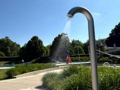 Eine silberne Außendusche verströmt feine Wasserstrahlen inmitten eines gepflegten Freibadgeländes mit viel Grün und freiem Himmel. Im Hintergrund erkennt man eine Wasserrutsche, Beckenanlagen und schattenspendende Bäume. Das Lichtspiel im Wassernebel und die klare Sommeratmosphäre erzeugen ein Gefühl von Erfrischung und Freizeitspaß.