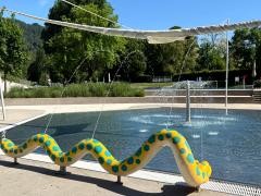 Das Bild zeigt ein flaches Kinderplanschbecken im Freien an einem sonnigen Tag. In der Mitte des Beckens sprudelt ein kleiner Springbrunnen aus einem senkrechten Metallrohr. Im Vordergrund befindet sich eine gewundene Figur in Schlangenform, die gelb ist und mit grünen Sechsecken bemalt wurde. Aus dieser Figur spritzen mehrere dünne Wasserstrahlen in das Becken. Darüber ist ein helles Sonnensegel aufgespannt, das teilweise Schatten spendet. Um das Becken herum sind grüne Pflanzen, Bäume und Rasenflächen zu sehen. Der Bereich wirkt ruhig, sauber und ist für Kinder zum Spielen geeignet.